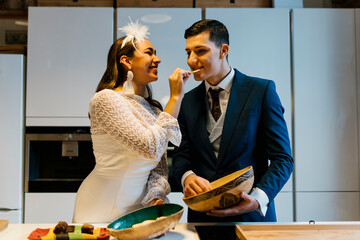 Smiling bride feeding potato chip to groom while standing in kitchen at home