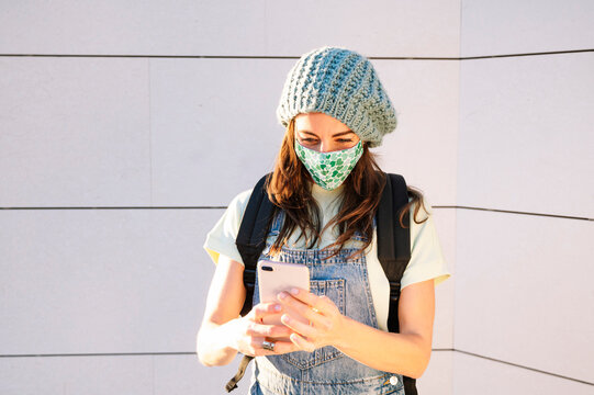 Mature woman using smart phone wearing protective face mask standing against wall on sunny day - Powered by Adobe