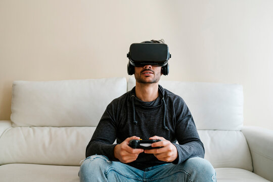 Young man wearing virtual reality simulator playing video game while sitting on sofa at home