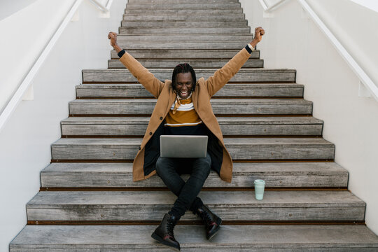Joyful African man using laptop while sitting on stairs