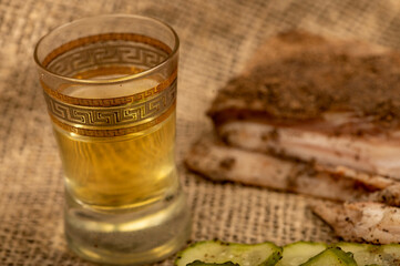 A piece of homemade bacon with pepper, a glass of homemade moonshine and chopped pickles close-up, selective focus.