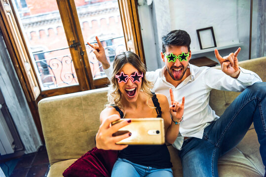 Playful young couple wearing comedy glasses and taking selfie on couch in living room at home