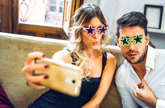 Playful young couple wearing comedy glasses and taking selfie on couch in living room at home