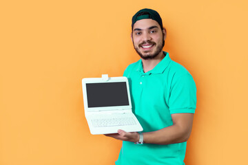 Young man with beard, T-shirt and green cap, showing to camera a tablet with keyboard, on yellow background.
