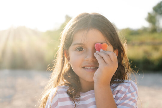 Close-up of cute smiling girl holding candy heart against face at park during sunset