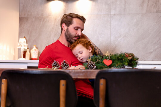 Father and son with Christmas wreath on table sleeping in kitchen at home