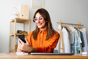 Female fashion designer working at home sitting at desk using smartphone