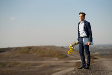 Mature businessman holding bunch of flowers and laptop on a disused mine tip