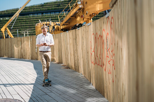 Businessman riding skateboard and using smartphone at construction site in the city