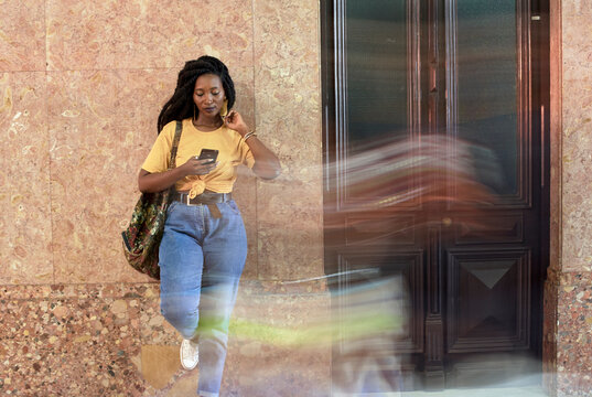 Young woman astanding at a wall checking her phone while person passing by