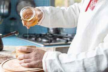 Obraz premium Ethnic linen shirt on the man. A man with a beard in the kitchen pours honey into a cup. Cooking dessert in a rustic kitchen.