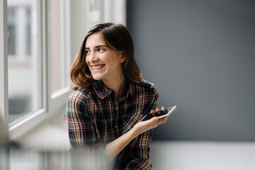 Portrait of laughing young woman with smartphone looking out of window