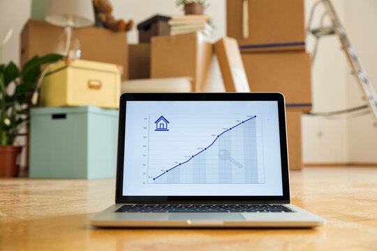 Rising line graph on laptop screen in front of cardboard boxes in an empty room in a new home