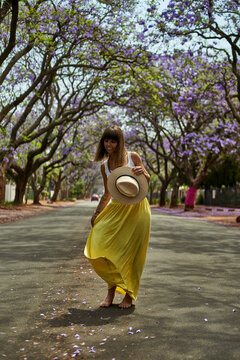 Woman dancing with a hat on her hands in the middle of a street full of jacaranda trees in bloom Pretoria, South Africa