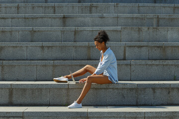 Positive multiracial young woman student in stylish clothes types on modern laptop sitting on large stone stairs on sunny summer day