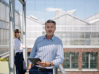Confident businessman holding digital tablet while standing with female coworker in greenhouse
