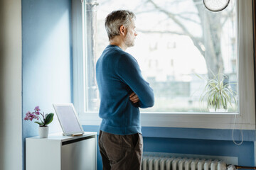 Pensive man standing in living room looking out of window