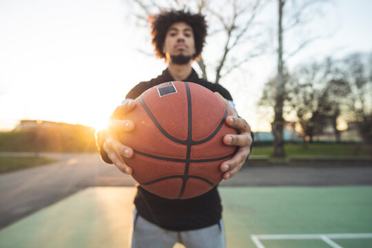 Young sportive man posing holding basketball on court at sunset