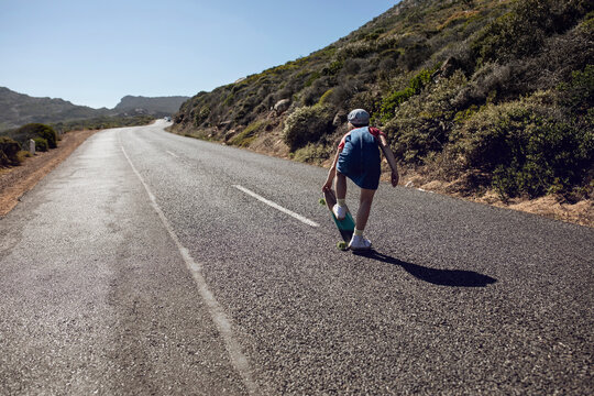 Back view of girl with skateboard standing on country road, Cape Town, Western Cape, South Africa