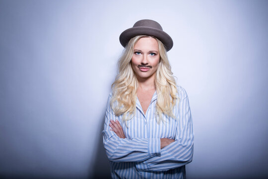 Portrait of blond young woman with fake moustache and hat crossing arms