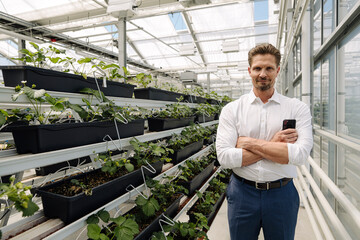 Confident male entrepreneur with arms crossed standing by plants in greenhouse