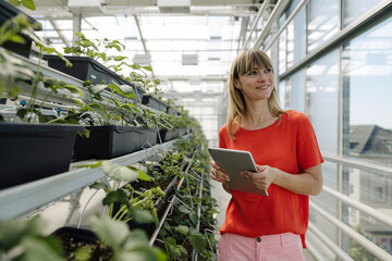 Smiling female entrepreneur with digital tablet looking away while standing by plants in nursery