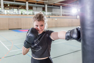 Smiling female boxer practising at punchbag in sports hall