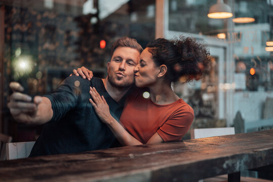 Young woman kissing man taking selfie through mobile phone while sitting at cafe