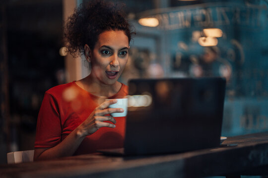 Young woman holding coffee cup while staring at laptop with mouth open in cafe