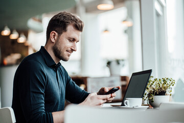 Mid adult businessman with laptop using smart phone while sitting at cafe