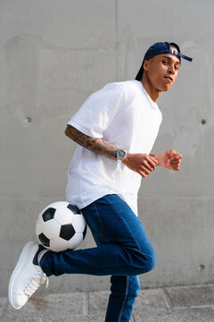 Portrait of tattooed young man balancing football in front of concrete wall
