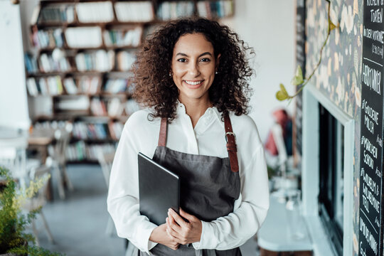 Smiling woman wearing apron holding book while standing at cafe