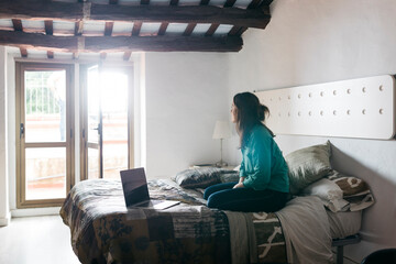 Young woman with laptop sitting on bed and looking out of the window