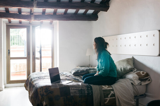 Young woman with laptop sitting on bed and looking out of the window