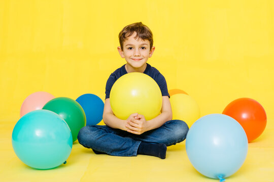 Portrait of smiling cute boy sitting with colorful balloons against yellow background