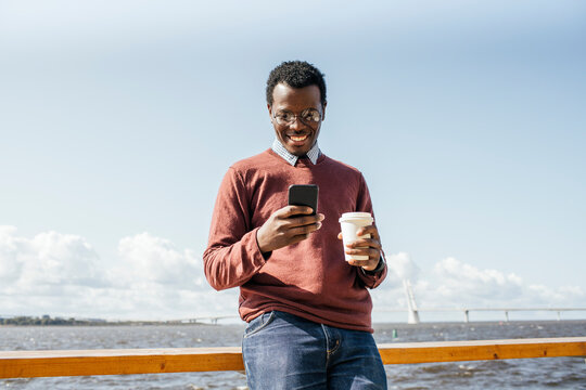 Young man standing at the sea, holding coffee, using smartphone