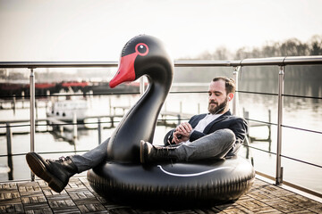 Hip businessman working on a houseboat, sitting a floating swan, using smartwatch