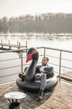 Hip businessman working on a houseboat, sitting a floating swan, using digital tablet