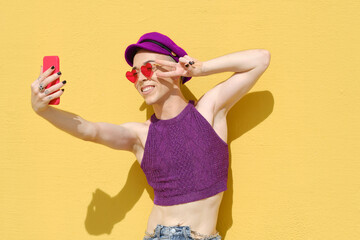 Non-binary young person taking selfies with a mobile phone while standing against a yellow wall.