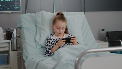 Sick little child resting in bed playing online video games using smartphone relaxing after...