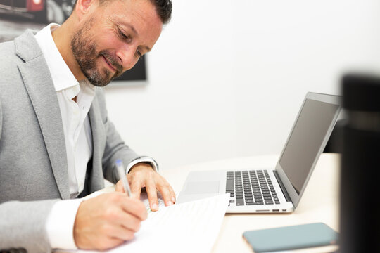 Male Entrepreneur With Laptop Writing On Paper At Desk In Office