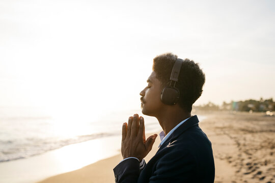 Afro man praying while wearing headphones at beach during sunny day