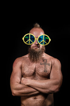 Shirtless male hipster wearing peace symbol novelty glasses standing with arms crossed against black background