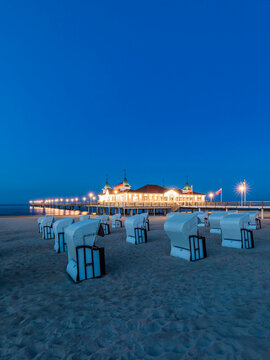 Germany, Mecklenburg-Western Pomerania, Heringsdorf, Hooded beach chairs on sandy coastal beach at dusk with illuminated pier in background