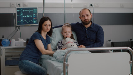 Portrait of sad family looking into camera while holding sick daughter hands waiting for medical therapy treatment against kid disease. Child lying in bed during recovery consultation in hospital ward