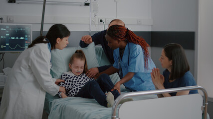 Hospitalized sick girl suffering pain attack while medical team holding her monitoring temperature during diagnosis examination in hospital ward. Pediatrician doctor trying to stop