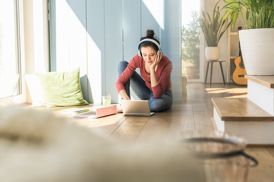 Woman with headphones sitting at the window at home using laptop