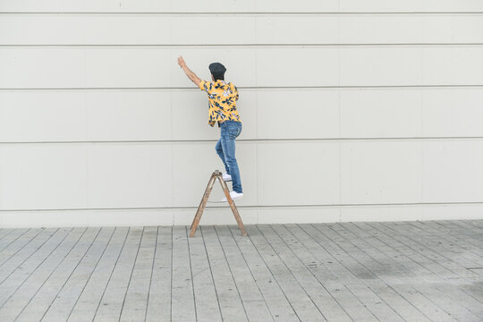 Young man wearing flat hat and aloa shirt, standing on step ladder, touching wall
