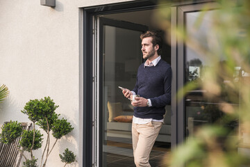 Young man leaning in door of his house, holding cup of coffee, using smartphone