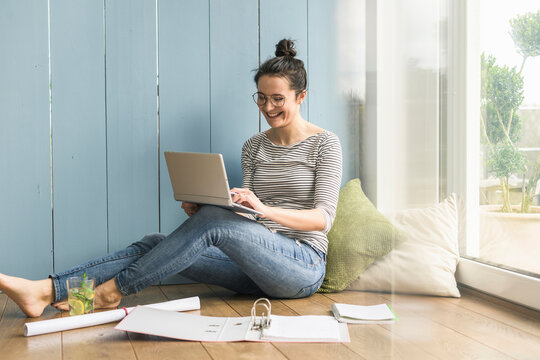 Smiling woman sitting at the window at home working with laptop and file folder